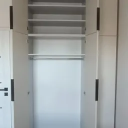 Jasna szafa z otwartymi drzwiami, widoczne półki i drążek na ubrania, minimalistyczny design z długimi, czarnymi uchwytami.