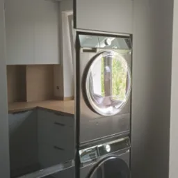 Szara zabudowa meblowa z pralką i suszarką w kolumnie, w tle fragment kuchni z jasnymi szafkami i drewnianym blatem. Minimalistyczny design, nowoczesne AGD.