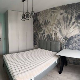 Meble Daniel Cebulski - Nowoczesna sypialnia z białą szafą na wymiar, łóżkiem z szarym zagłówkiem i biurkiem. Na ścianie dekoracyjna tapeta w motywy roślinne, a nad szafą lampy wiszące.