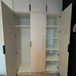 Meble Daniel Cebulski - Jasna szafa wnękowa z otwartymi drzwiami, widoczne półki i minimalistyczne czarne uchwyty, na podłodze panele imitujące drewno, czarna ściana w tle.