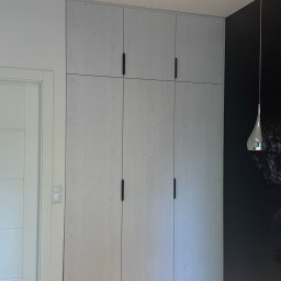 Meble Daniel Cebulski - Jasnoszara szafa wnękowa z czarnymi uchwytami, dopasowana do wysokości pomieszczenia, obok drzwi i ciemnej ściany z lampą wiszącą.