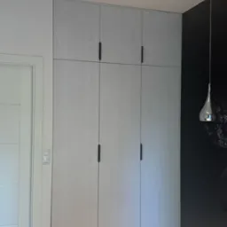 Jasnoszara szafa wnękowa z czarnymi uchwytami, dopasowana do wysokości pomieszczenia, obok drzwi i ciemnej ściany z lampą wiszącą.