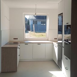 Meble Daniel Cebulski - Jasna kuchnia z białymi frontami i drewnianymi akcentami, widok na ogród przez okno. W tle dom z numerem 16. Nowoczesny design, minimalistyczne uchwyty.