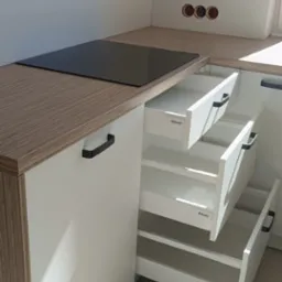 Jasna kuchnia z blatem imitującym drewno. Otwarta szafka z trzema szufladami Blum. Widoczna płyta indukcyjna i gniazdka elektryczne w ścianie.