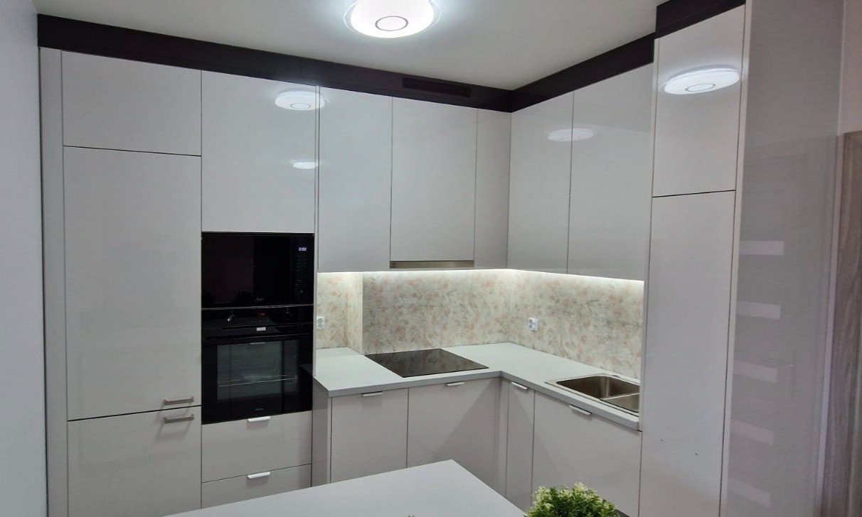 Nowoczesna, jasna kuchnia z białymi, lakierowanymi frontami, czarnym piekarnikiem i płytą indukcyjną, oświetleniem LED pod szafkami wiszącymi i blatem w odcieniach szarości.