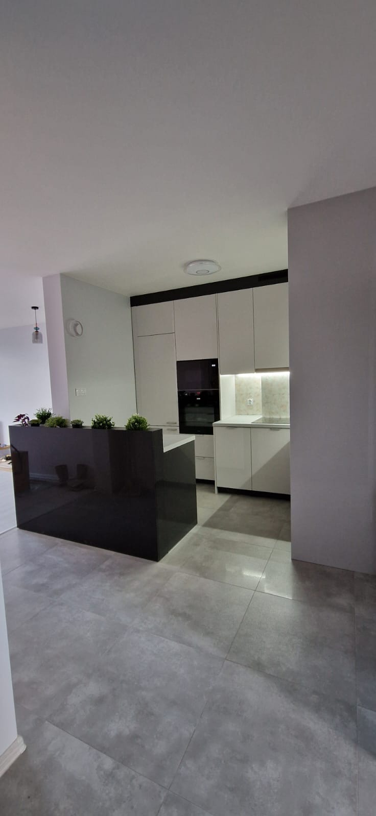 Nowoczesna kuchnia z czarnym, lśniącym półwyspem i białymi szafkami, oświetlenie LED pod szafkami górnymi i rośliny doniczkowe na blacie półwyspu.