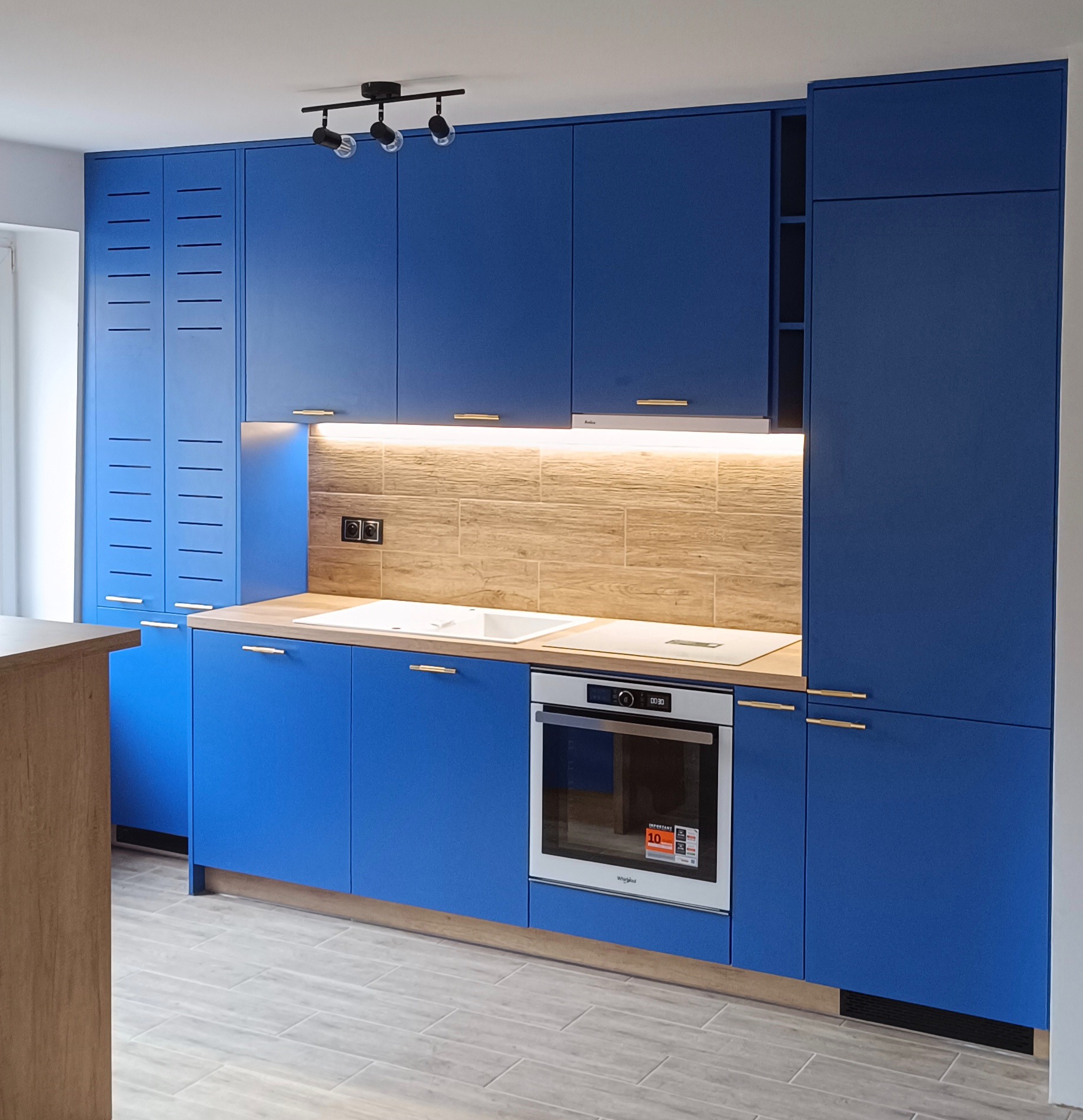 Niebieska zabudowa kuchenna z drewnianym blatem, białym zlewem i płytą indukcyjną, piekarnik Whirlpool, oświetlenie podszafkowe i sufitowe, płytki imitujące drewno na ścianie.