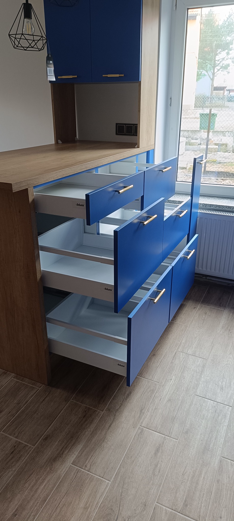 Niebieska zabudowa kuchenna z drewnianym blatem, otwarte szuflady z systemem Blum, widok na okno i grzejnik.