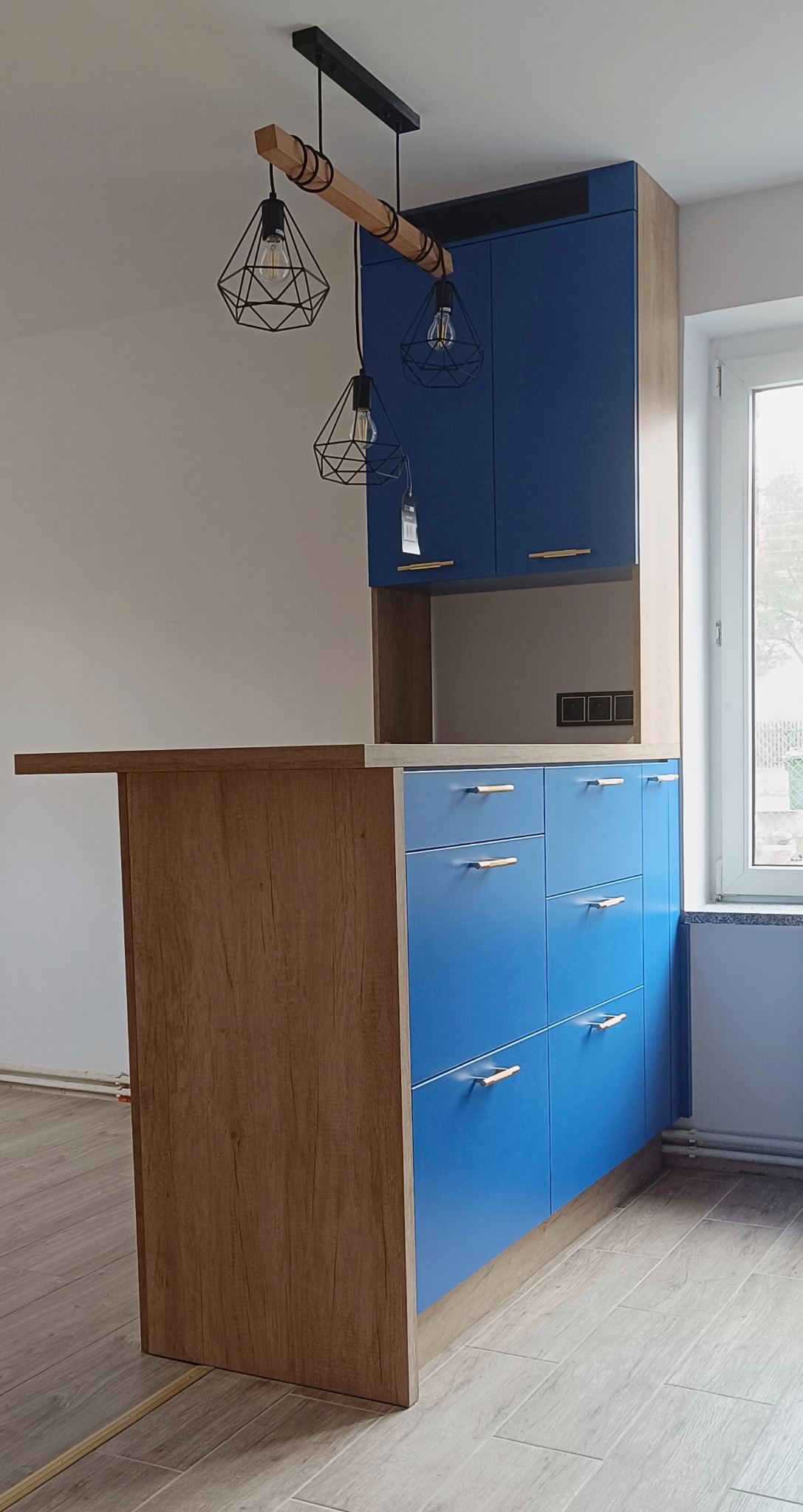 Niebieskie szafki kuchenne z drewnianymi akcentami i lampami wiszącymi w stylu industrialnym, widok na okno w tle.