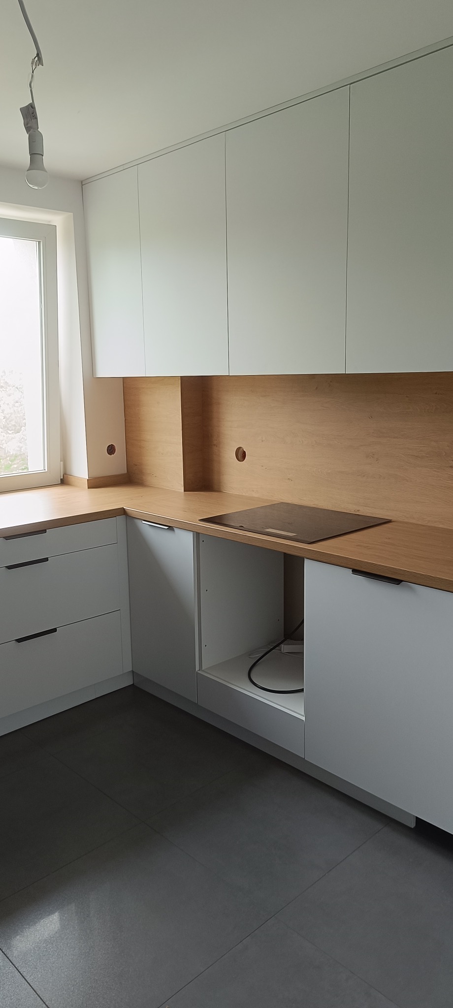 Jasna kuchnia z białymi szafkami i drewnianym blatem. Widoczne otwory na gniazdka. Minimalistyczny styl, szare płytki podłogowe, okno po lewej stronie.