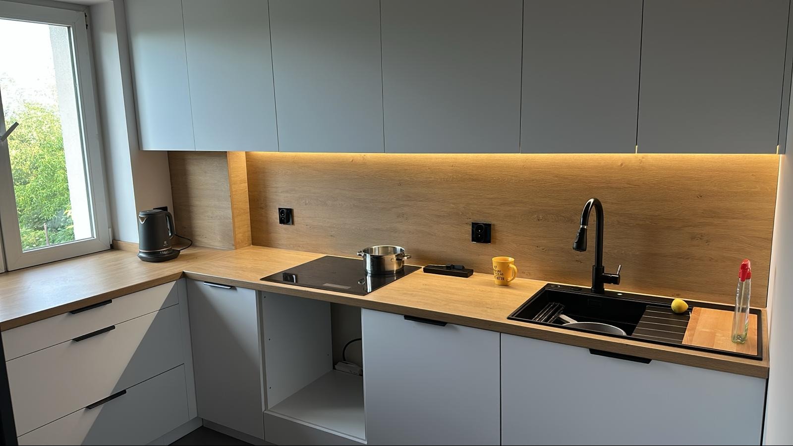 Nowoczesna kuchnia z szarymi szafkami, drewnianym blatem i oświetleniem LED. Na blacie czajnik, płyta indukcyjna, zlew z czarną armaturą i żółty kubek.