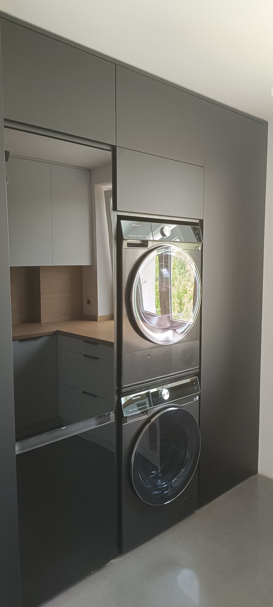 Szara zabudowa meblowa z pralką i suszarką w kolumnie, w tle fragment kuchni z jasnymi szafkami i drewnianym blatem. Minimalistyczny design, nowoczesne AGD.