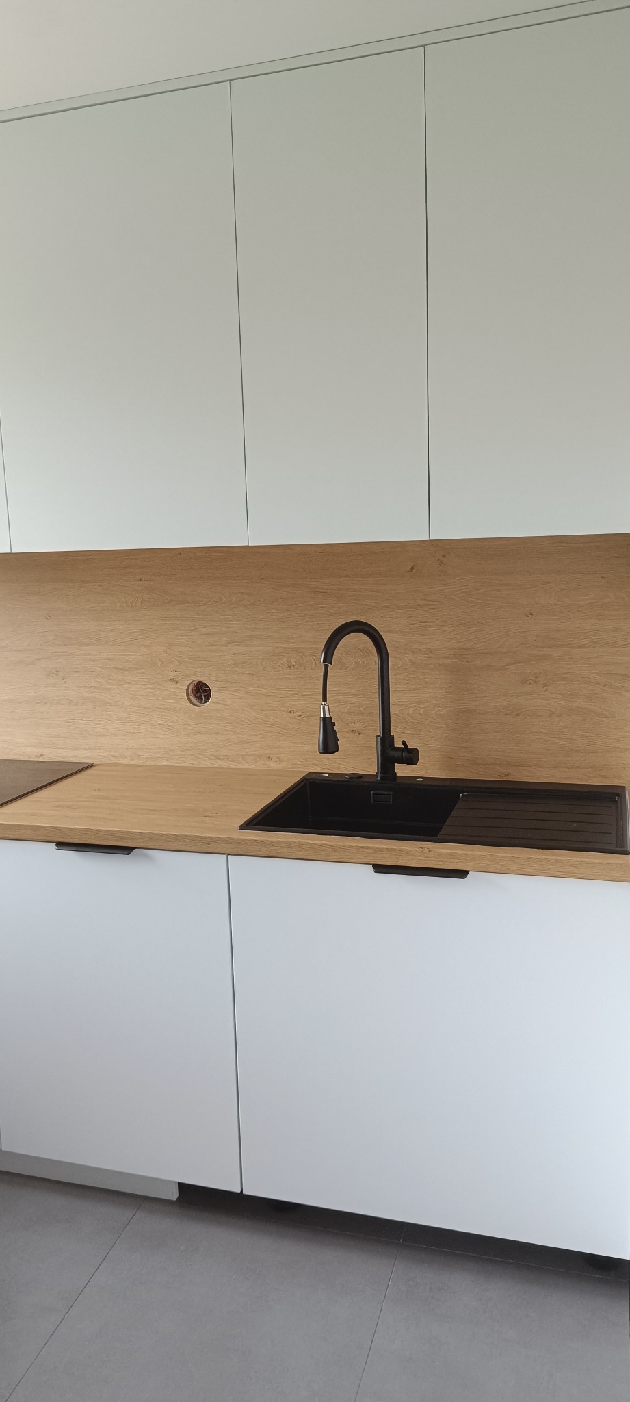 Minimalistyczna kuchnia: białe szafki, drewniany blat z czarnym zlewem i baterią. Widoczny otwór na kabel w blacie. Nowoczesny design, jasne wnętrze.