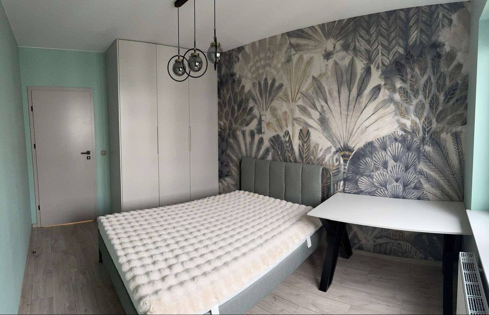Nowoczesna sypialnia z białą szafą na wymiar, łóżkiem z szarym zagłówkiem i biurkiem. Na ścianie dekoracyjna tapeta w motywy roślinne, a nad szafą lampy wiszące.