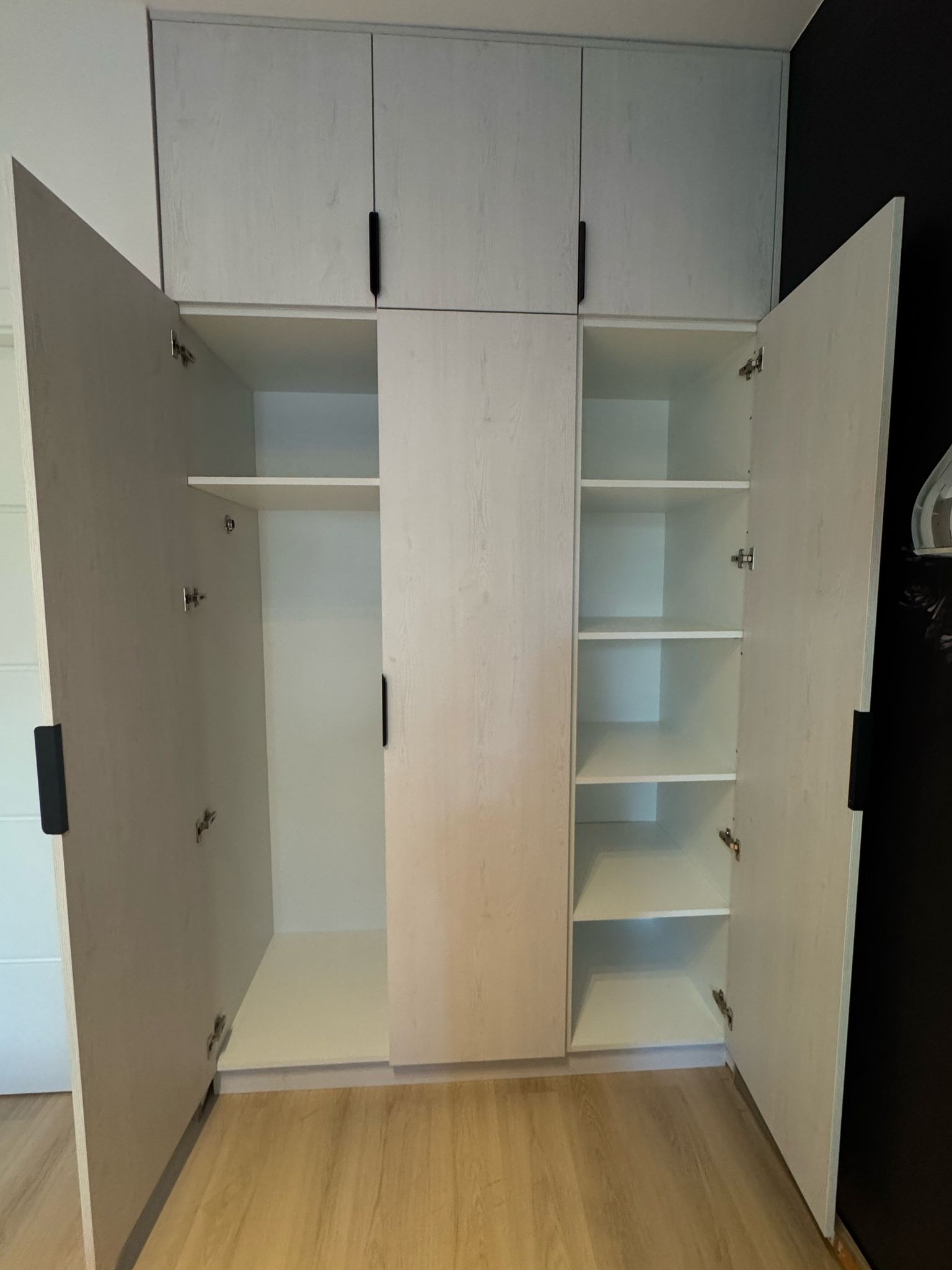 Jasna szafa wnękowa z otwartymi drzwiami, widoczne półki i minimalistyczne czarne uchwyty, na podłodze panele imitujące drewno, czarna ściana w tle.