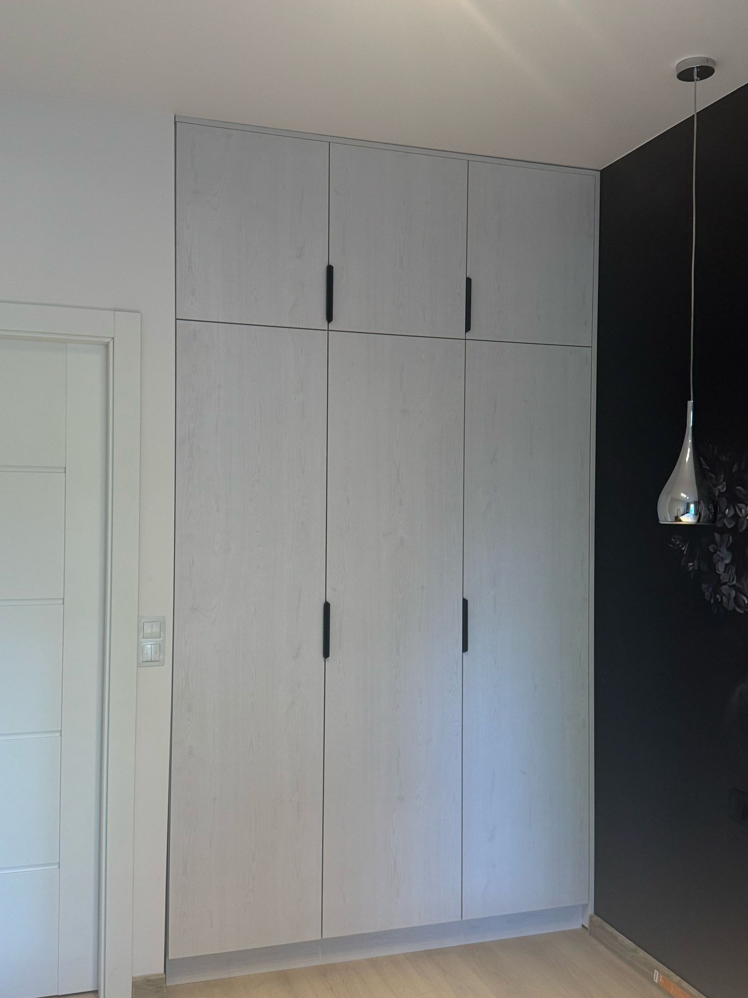 Jasnoszara szafa wnękowa z czarnymi uchwytami, dopasowana do wysokości pomieszczenia, obok drzwi i ciemnej ściany z lampą wiszącą.