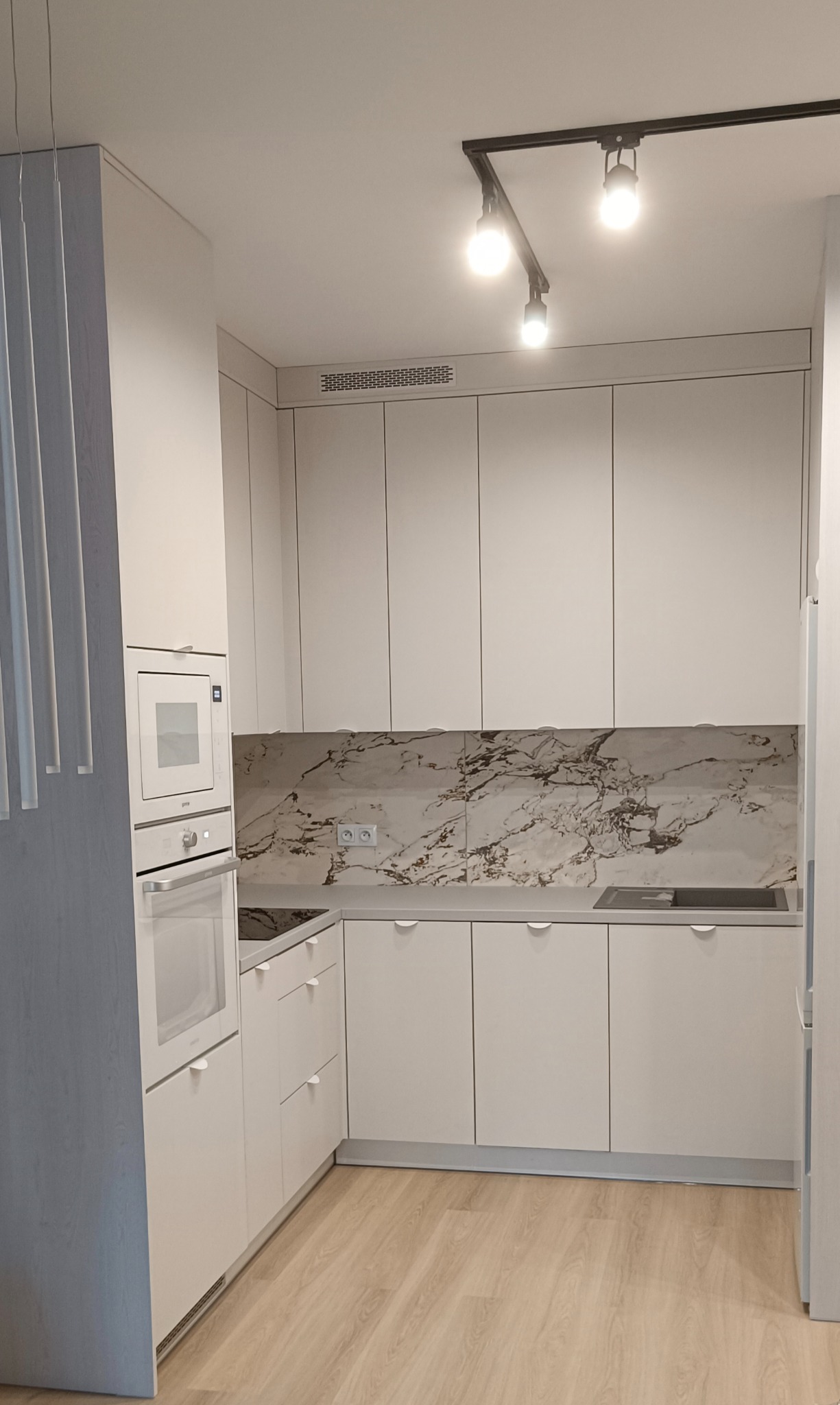 Jasna kuchnia z białymi frontami i marmurowym backsplashiem. Oświetlenie punktowe na czarnej szynie. Minimalistyczne uchwyty. Nowoczesny piekarnik i płyta grzewcza.