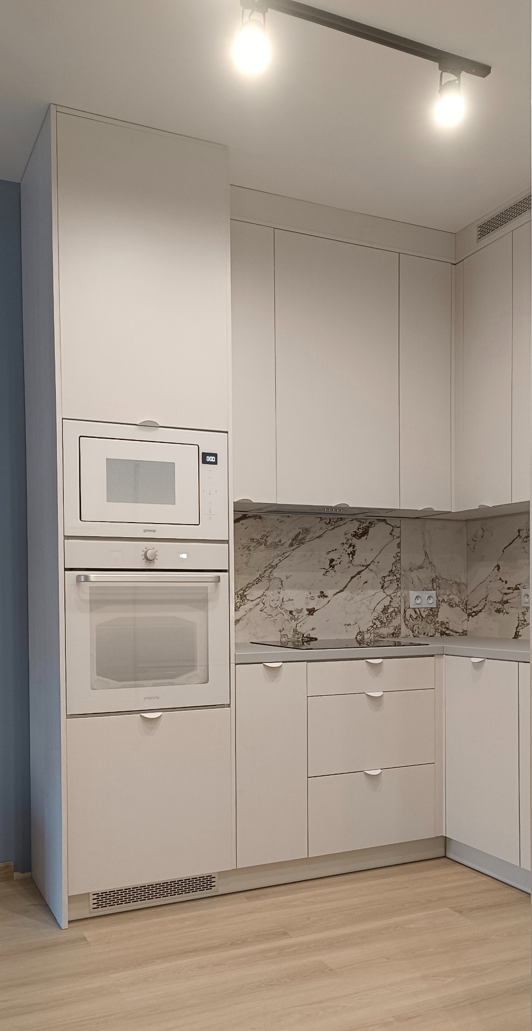 Jasna kuchnia na wymiar z zabudową piekarnika i mikrofalówki Gorenje, marmurowy backsplash. Oświetlenie szynowe i drewniana podłoga.