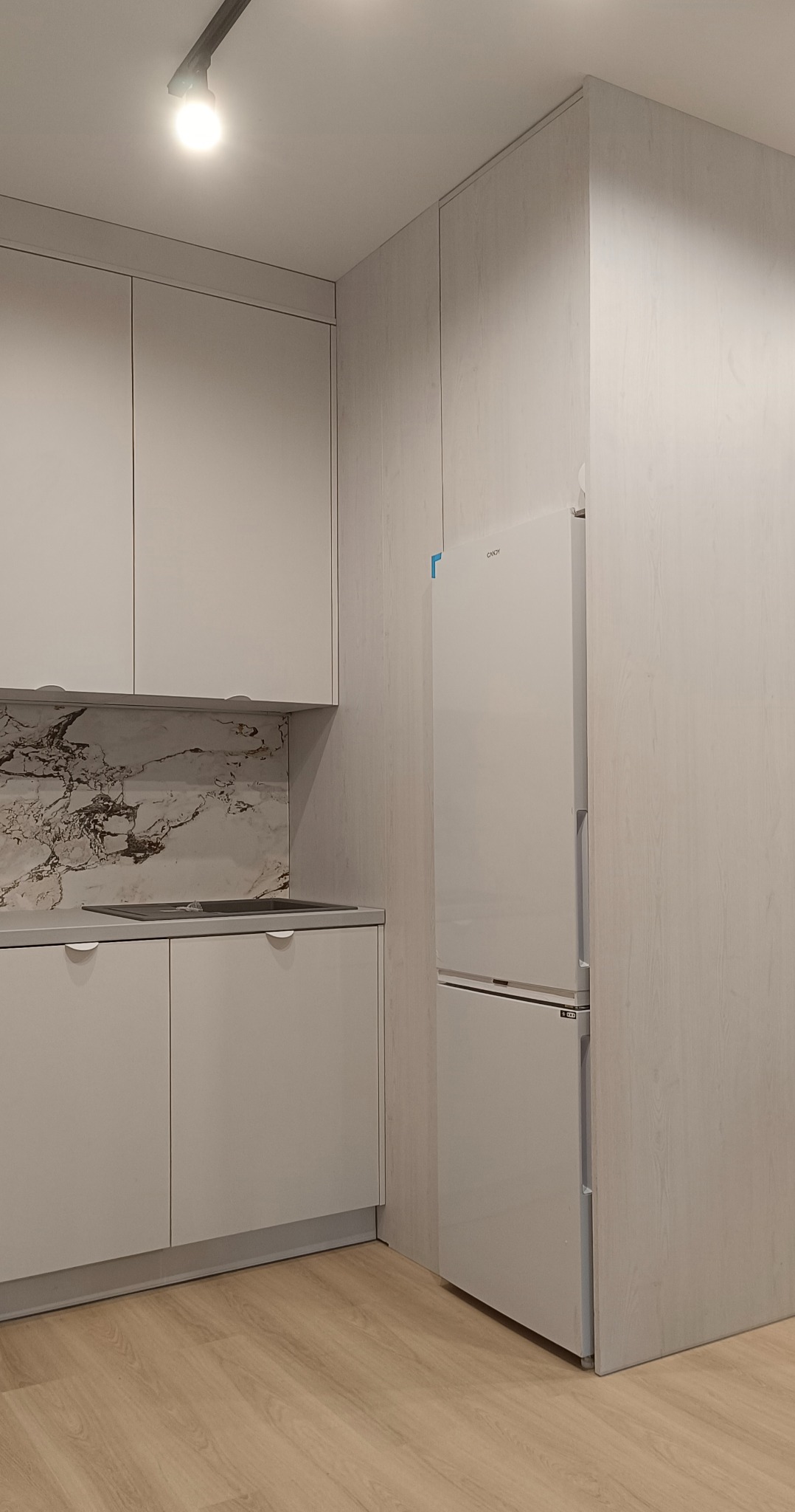 Jasna kuchnia z frontami bez uchwytów, marmurowym panelem nad blatem i lodówką w zabudowie. Minimalistyczny design i oświetlenie punktowe.