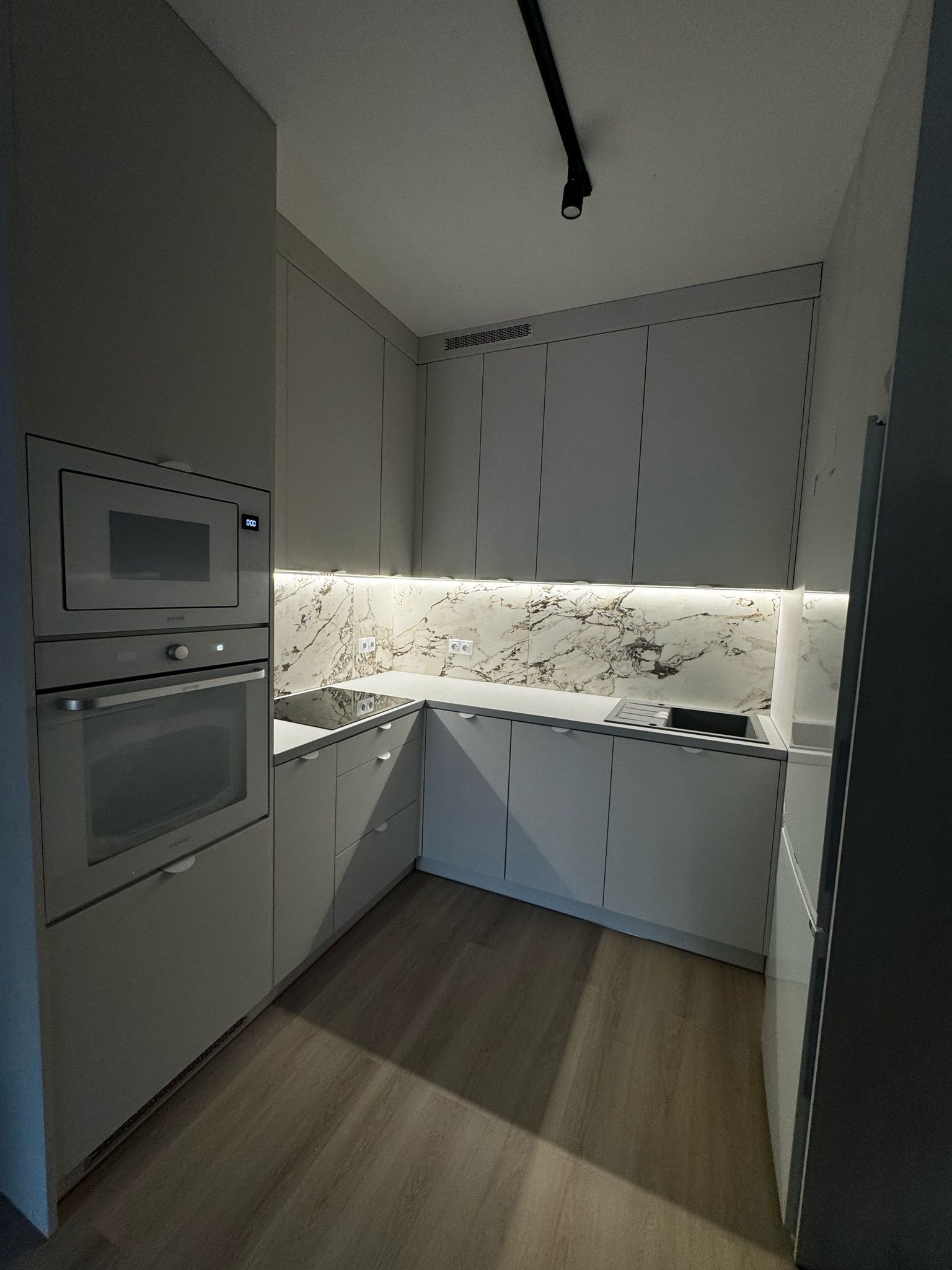 Nowoczesna, jasna kuchnia z marmurowym backsplashiem i oświetleniem LED. Minimalistyczny design, zabudowa kuchenna z piekarnikiem i mikrofalówką.