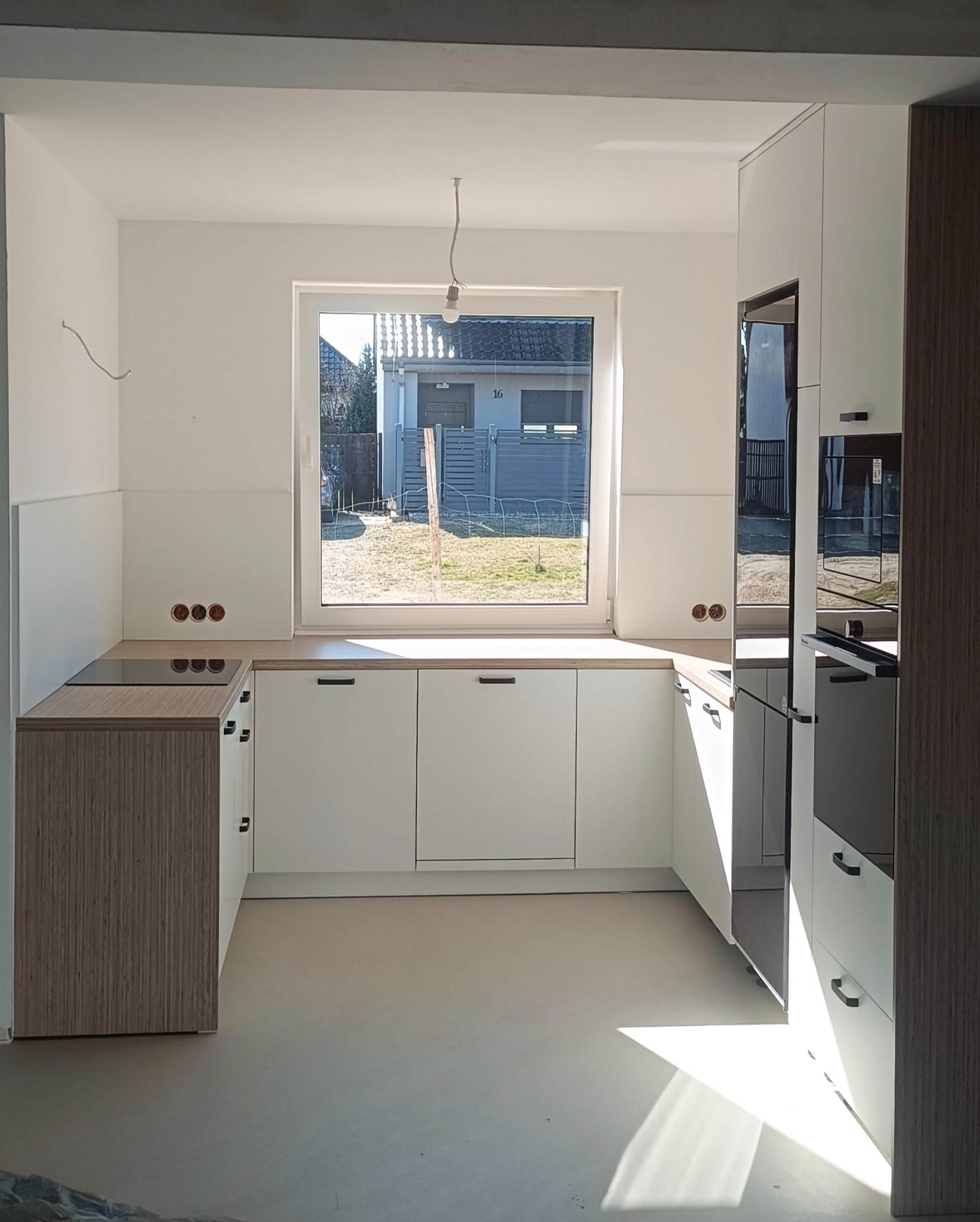 Jasna kuchnia z białymi frontami i drewnianymi akcentami, widok na ogród przez okno. W tle dom z numerem 16. Nowoczesny design, minimalistyczne uchwyty.