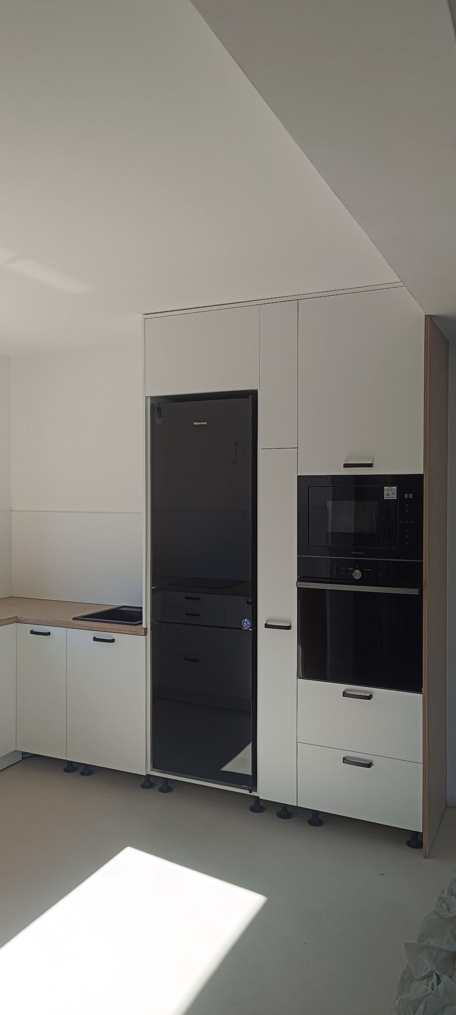 Nowoczesna kuchnia z białymi frontami, czarnym sprzętem AGD (lodówka Hisense, piekarnik i mikrofalówka). Minimalistyczny design, jasne wnętrze z drewnianym blatem.