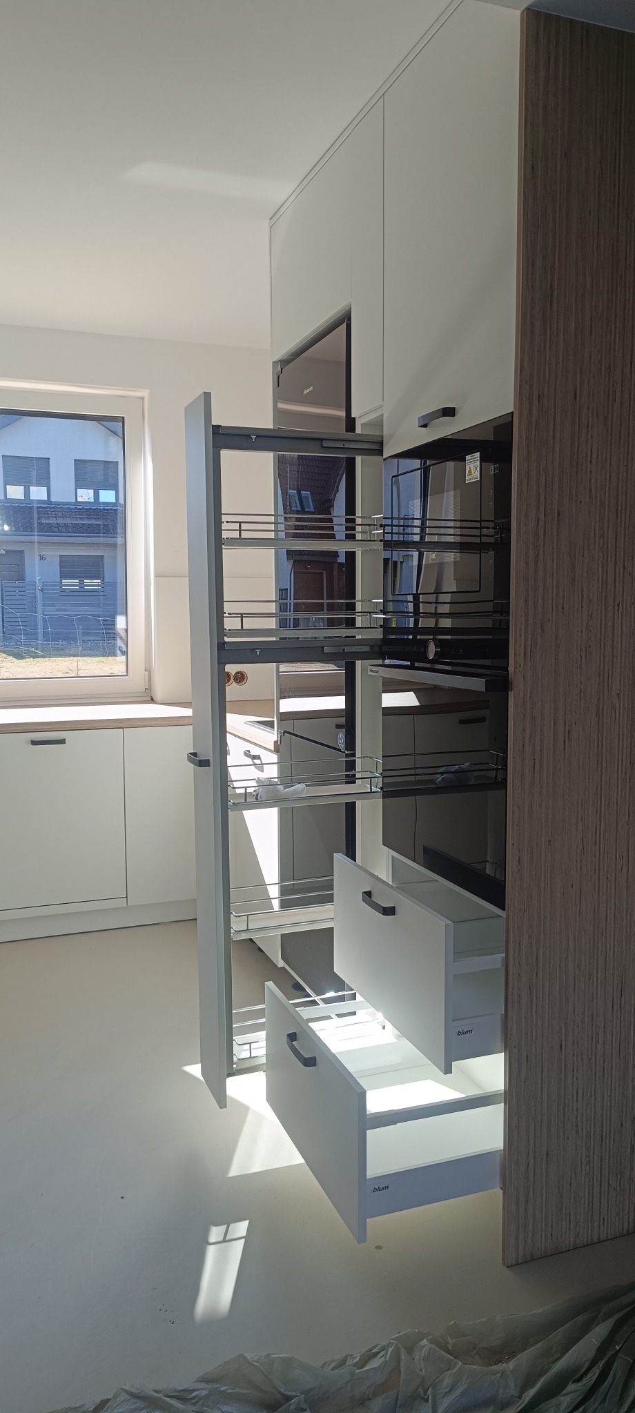 Wysuwana szafka cargo z metalowymi koszami i szufladami w nowoczesnej, białej zabudowie kuchennej z drewnopodobnym bokiem. Widok na okno i fragment zabudowy.