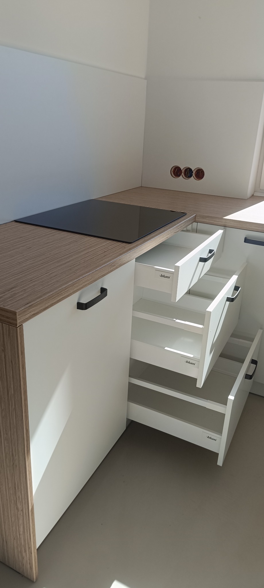 Jasna kuchnia z blatem imitującym drewno. Otwarta szafka z trzema szufladami Blum. Widoczna płyta indukcyjna i gniazdka elektryczne w ścianie.