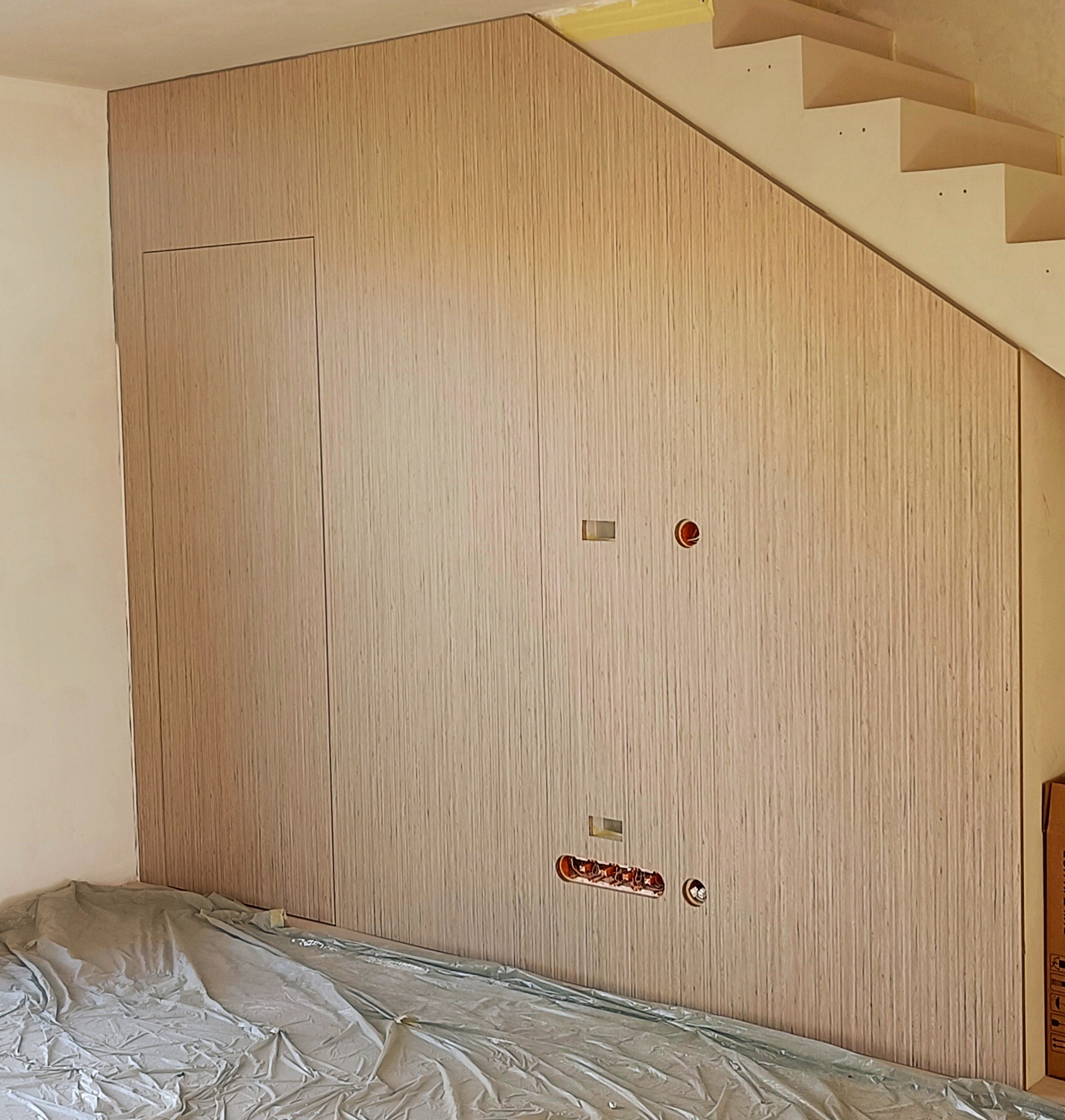 Szafa wnękowa pod schodami, wykończona okładziną drewnopodobną z widocznymi otworami na gniazdka elektryczne, zabezpieczona folią malarską.