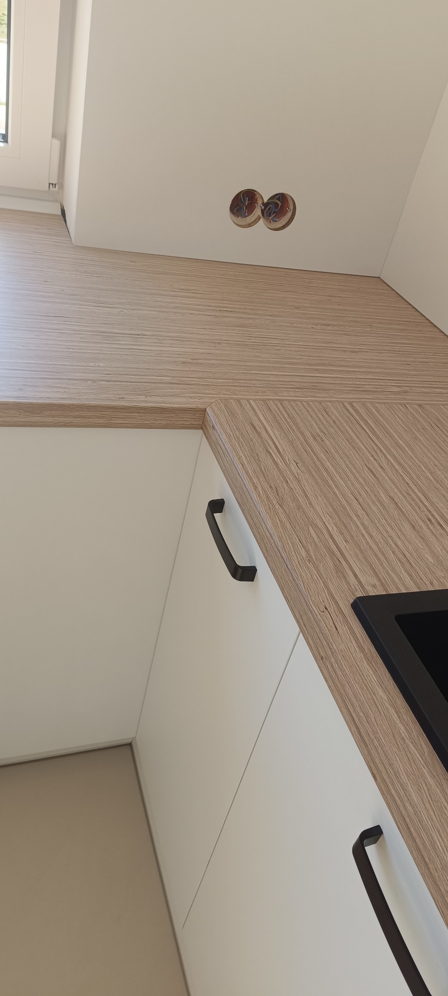 Fragment nowoczesnej, jasnej kuchni z drewnianym blatem i czarnym, minimalistycznym uchwytem szafki. Widoczne gniazdka elektryczne w ścianie.