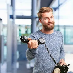 Uśmiechnięty mężczyzna z brodą, trzymający w wyciągniętej ręce czarny telefon stacjonarny w stylu retro, z kablem zwisającym w dół, na rozmytym tle biurowym.