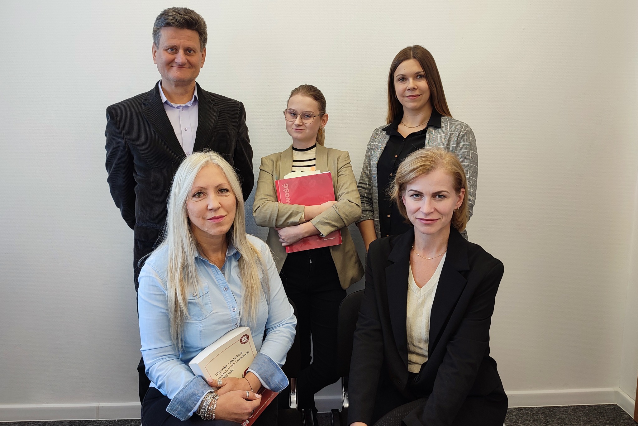 Zespół pięciu osób w biurowym otoczeniu, kobiety trzymają książki, uśmiechają się do kamery.