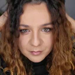 Portret kobiety z zielonymi oczami, delikatnym błyszczącym makijażem ust i brązowymi kręconymi włosami z ciemnymi odrostami, trzymającej ręce za głową.