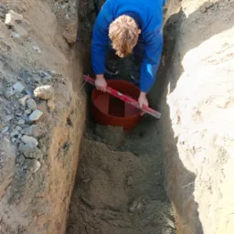 Pracownik w niebieskiej bluzie poziomuje czerwoną studzienkę kanalizacyjną w wykopie o pionowych ścianach, używając poziomicy.