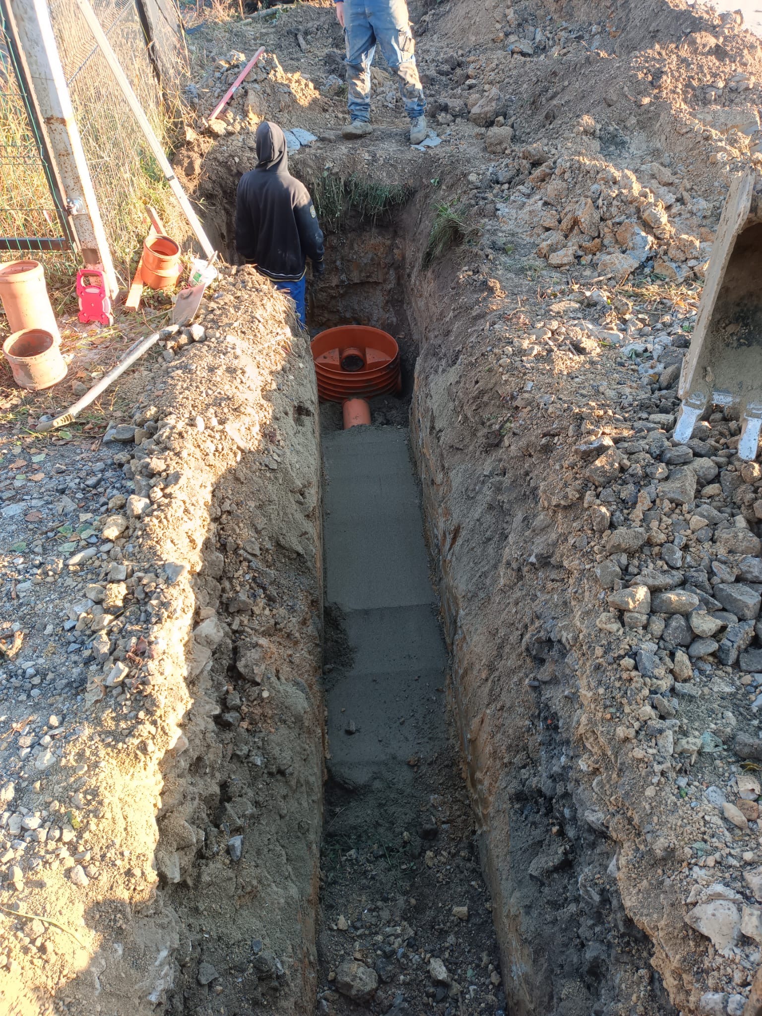 Wykop liniowy z widocznymi elementami instalacji kanalizacyjnej w trakcie montażu, w tle pracownik w bluzie z kapturem i fragment koparki.