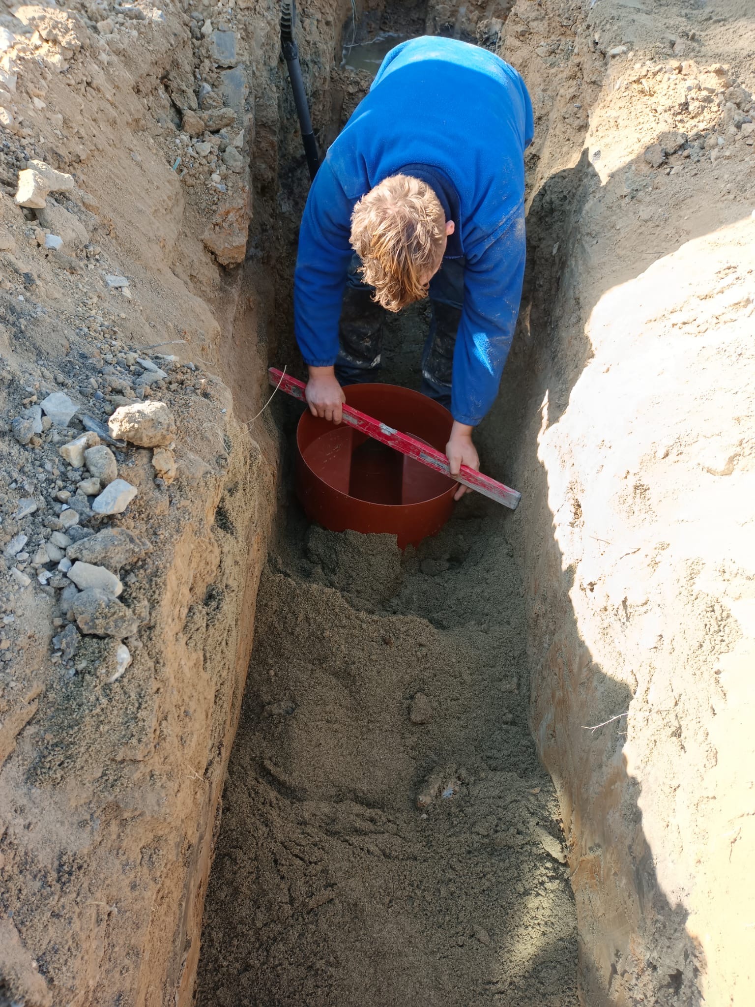 Pracownik w niebieskiej bluzie poziomuje czerwoną studzienkę kanalizacyjną w wykopie o pionowych ścianach, używając poziomicy.