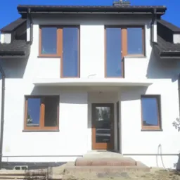 Biały dom jednorodzinny z brązowymi oknami i drzwiami, po remoncie elewacji, widoczne materiały budowlane na trawniku.