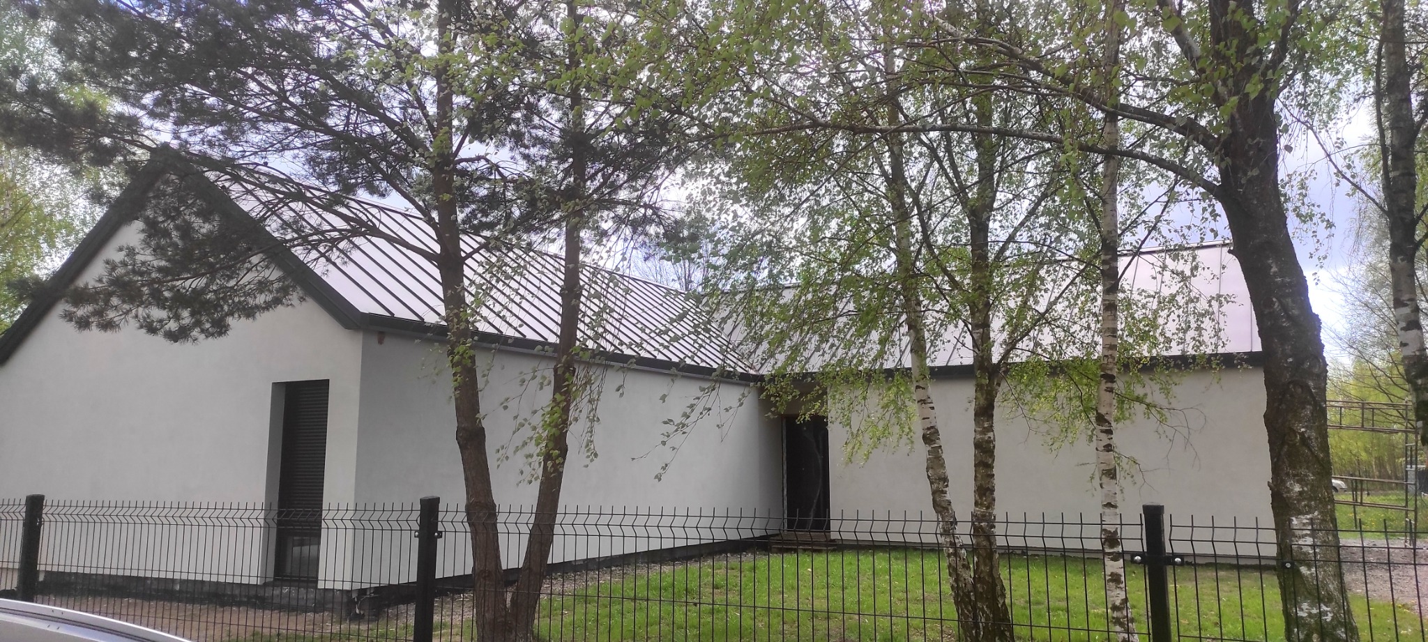 Biały budynek z ciemnym dachem i czarną stolarką, widziany zza metalowego ogrodzenia i drzew, wczesna wiosna.