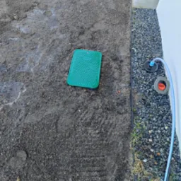 Instalacja systemu nawadniania: zielona skrzynka zaworowa w ziemi, obok rura i zraszacze na kamienistym podłożu przy ścianie budynku. Widok z góry.