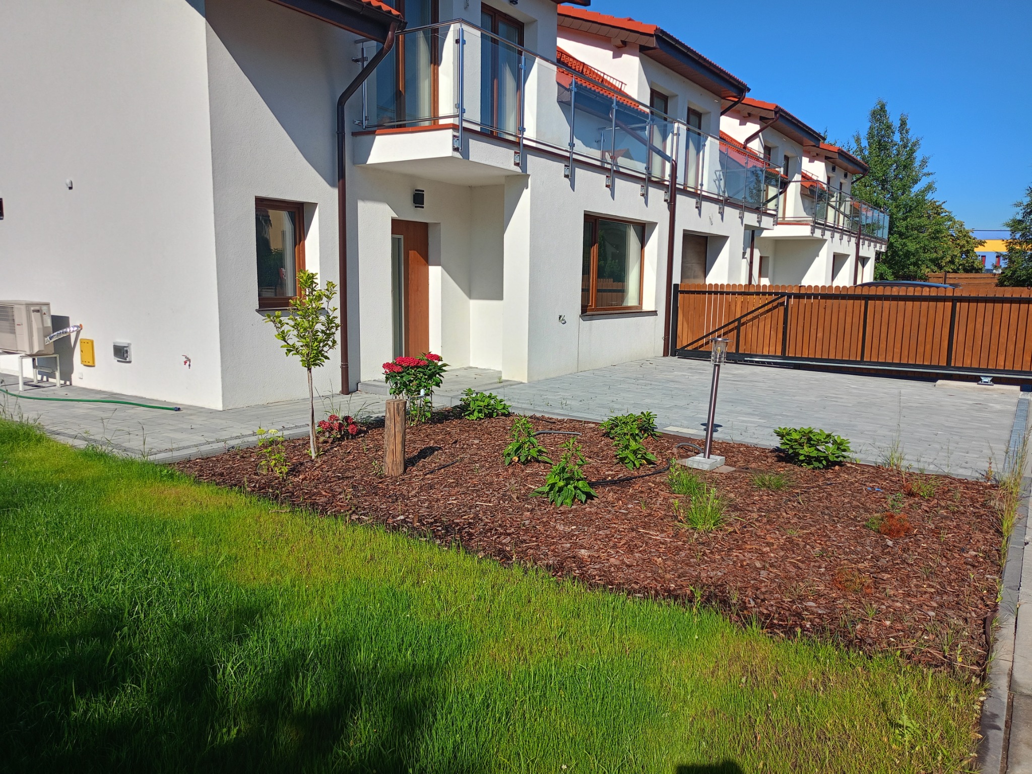 Świeżo założony ogród przed nowoczesnym domem szeregowym z białymi elewacjami, widoczny trawnik, rabata wyłożona korą, niskie krzewy, młode drzewko i kwiaty w donicy przy wejściu.