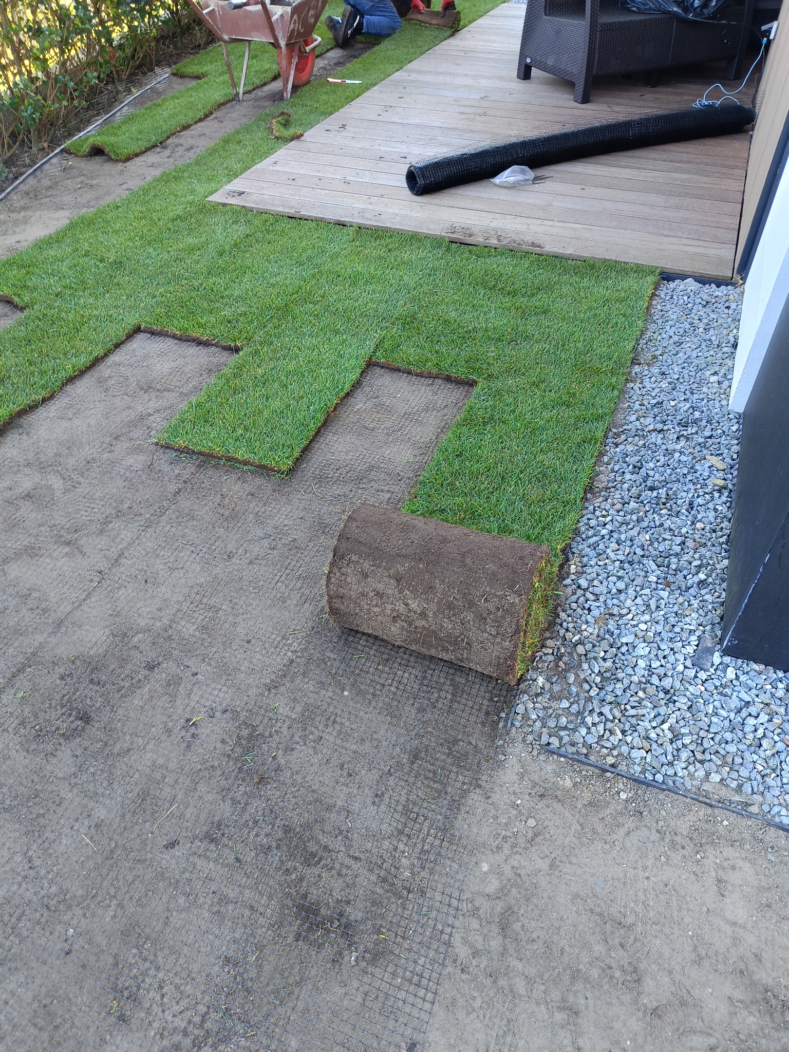 Układanie trawy z rolki w ogrodzie, widok z góry. Częściowo ułożona trawa, rolka trawy leży obok kamyków. W tle taczka i fragment drewnianego tarasu.