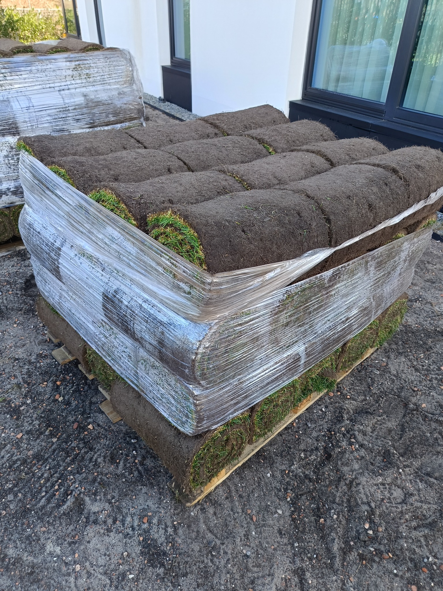 Paleta z rolkami trawy, zabezpieczona folią, gotowa do transportu. Widoczne warstwy darni z zieloną trawą i ciemną ziemią. W tle fragment budynku z oknem.