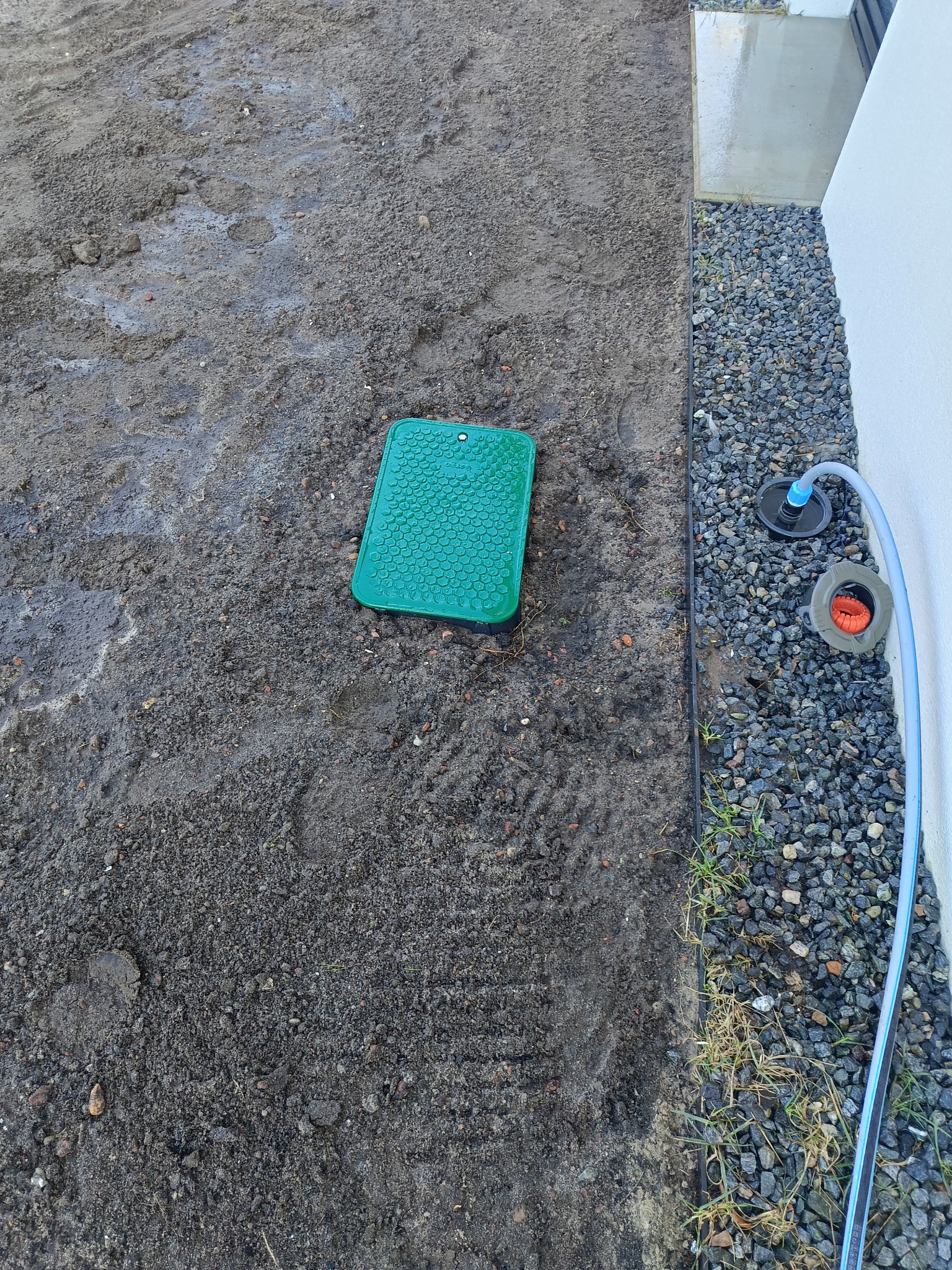 Instalacja systemu nawadniania: zielona skrzynka zaworowa w ziemi, obok rura i zraszacze na kamienistym podłożu przy ścianie budynku. Widok z góry.