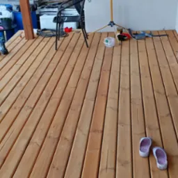 Świeżo położony drewniany taras z widocznymi narzędziami, lampą budowlaną, parą klapek i donicą przy oknie.