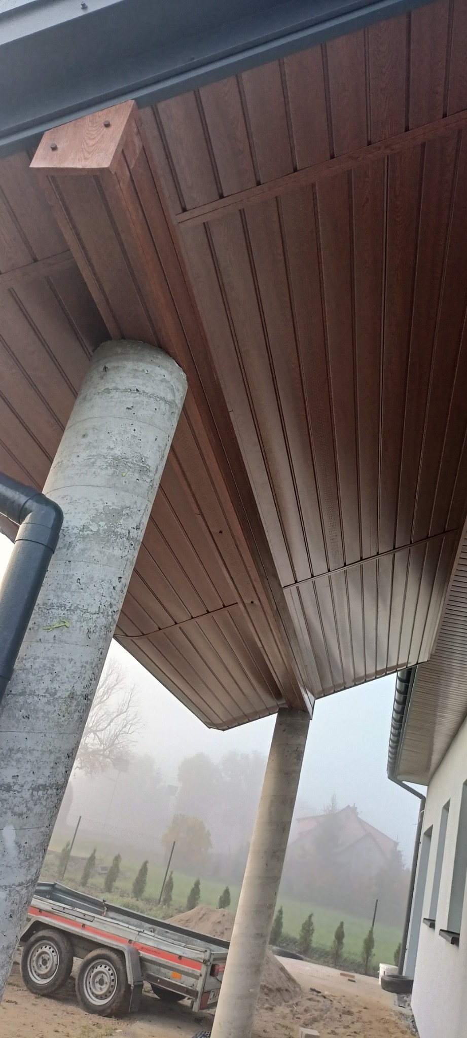 Podbitka dachowa z brązowych paneli drewnopodobnych, wsparta na betonowych kolumnach, widok z dołu na tle mglistego krajobrazu i zaparkowanej przyczepy.