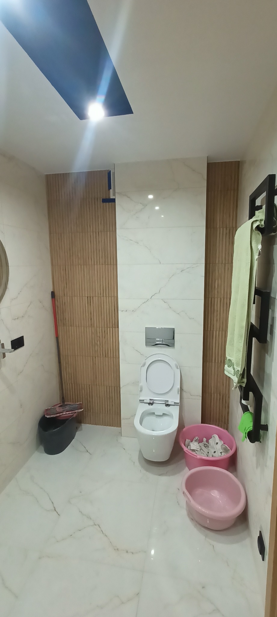 Wykończona łazienka z marmurowymi płytkami, drewnianymi panelami ściennymi, i białą toaletą z otwartą klapą, widoczne akcesoria łazienkowe i wiaderka z zabrudzeniami po remoncie.