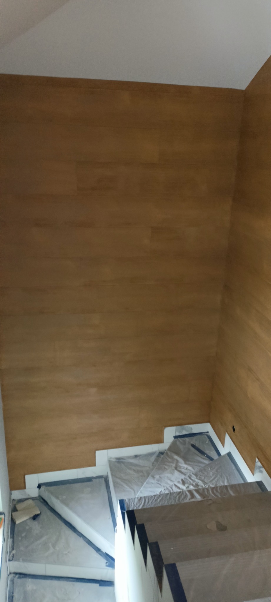Ściana wykończona panelami drewnopodobnymi w odcieniach brązu, schody zabezpieczone folią i taśmą malarską podczas prac wykończeniowych.
