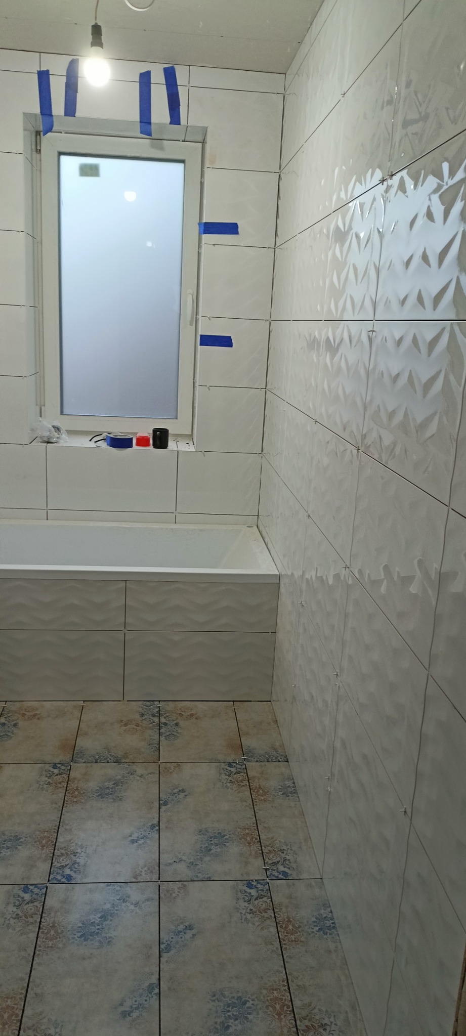 Białe, trójwymiarowe płytki ścienne w trakcie montażu w łazience, z widocznymi krzyżykami dystansowymi i niebieską taśmą malarską wokół okna; podłoga wyłożona płytkami w stylu vintage.