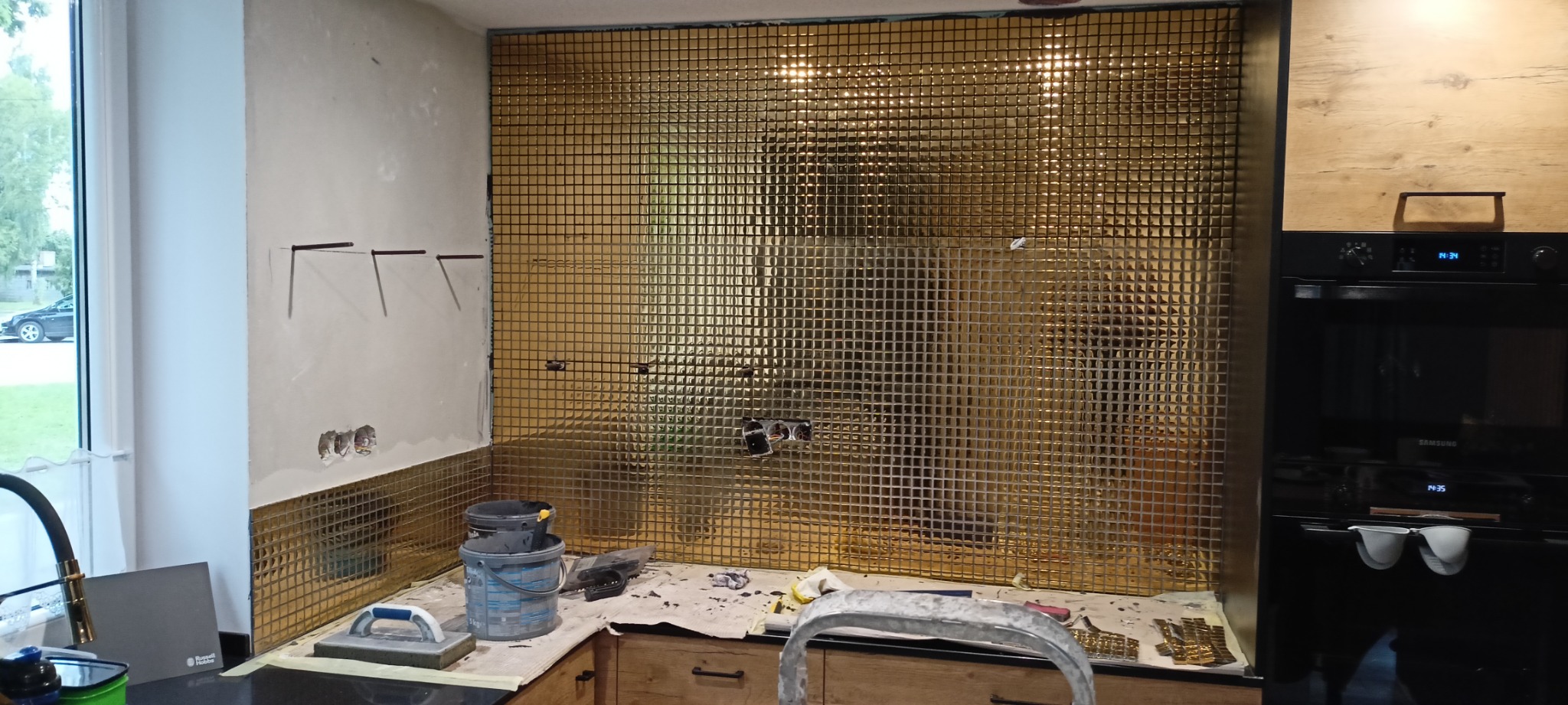 Ściana w kuchni pokryta złotą mozaiką w trakcie prac, widoczne narzędzia glazurnicze, wiaderka z klejem, fragment ściany z przygotowanymi otworami na gniazdka elektryczne i wystającymi wspornikami,...