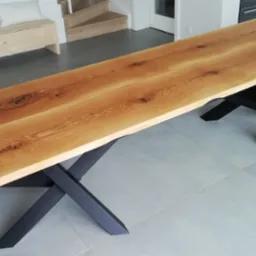 Długi blat drewniany o naturalnym wykończeniu z widocznymi słojami i sękami, osadzony na czarnych, metalowych nogach w kształcie litery X, ustawiony w nowoczesnym wnętrzu z szarymi płytkami...